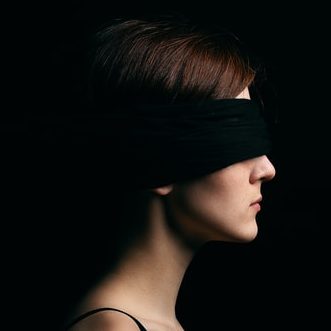 Una mujer joven de perfil, con un pañuelo negro que le tapa los ojos y el fondo también negro.